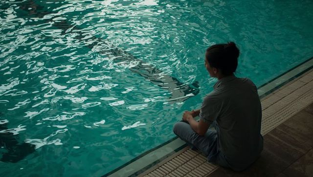Trainer sitting at pool edge watching dolphin shadow in turquoise water with ripples
