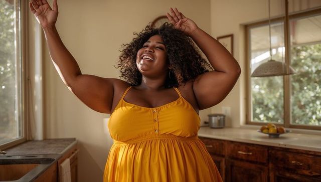 Curvy black woman dancing and smiling in sunlit rustic kitchen wearing mustard sundress