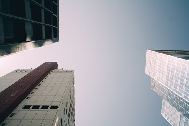 Depicts an urban scene with modern skyscrapers reaching up to a clear, cloudless sky. These buildings, with their mix of glass and concrete, reflect modern architectural trends. Suitable for backgrounds, city-related content, architecture topics, business, and urban lifestyle themes. Effective for illustrating concepts of modern living, corporate environments, and metropolitan skylines.