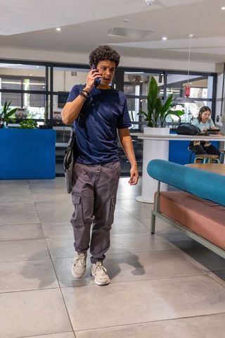 Young man walking through coworking lounge talking on phone carrying messenger bag