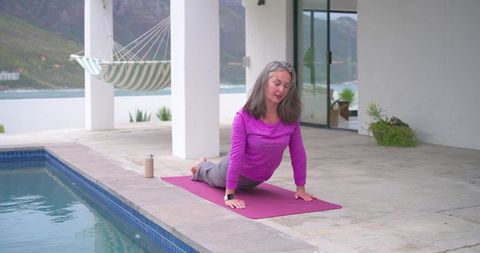 Senior woman practicing gentle backbend on magenta mat by pool with ocean view