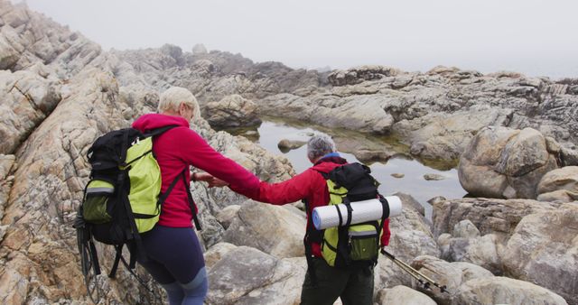 Senior hiker couple with backpacks and trekking poles holding each others hands while climbing rocks in the mountains. trekking, hiking, nature, activity, exploration, adventure concept.