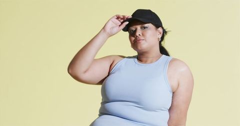 Confident woman tipping cap in activewear against pastel background
