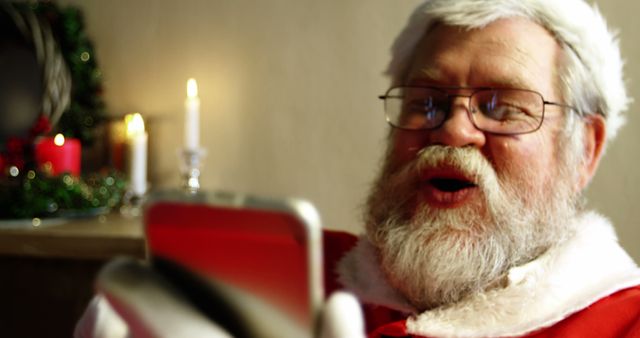 A Caucasian man dressed as Santa Claus is reading from a tablet, with copy space. His expression suggests he is engaged in a cheerful activity, reading messages or a list, in a festive environment.