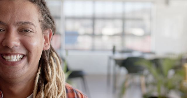 Smiling young man with dreadlocks in casual attire showing positive emotion and happiness in a bright office workspace. Ideal for use in advertisements, corporate culture promotion, or any content showcasing a positive and inclusive work environment.