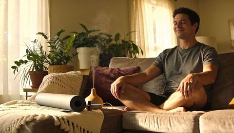 Man meditating on sunlit sofa with diffuser and yoga mat for home wellness