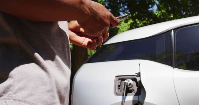 Midsection of african american senior man charging electric car and using smartphone on a sunny day. at home preparing eco-friendly car for journey.