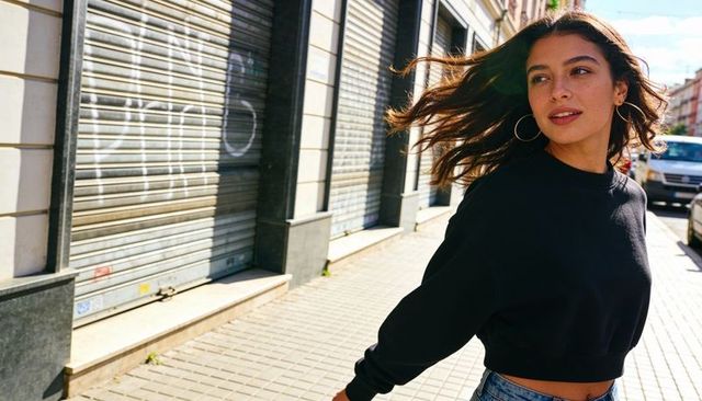 Windblown young woman walking in black sweatshirt and denim on sunny urban street