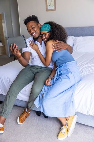 Couple hugging and smiling while using tablet on bed edge in cozy modern bedroom