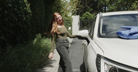 Redhead woman touching suv side mirror smiling on leafy suburban driveway with blue bag