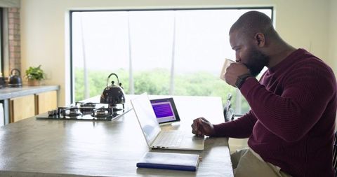 African American man working remotely at kitchen island, sipping coffee and using laptop