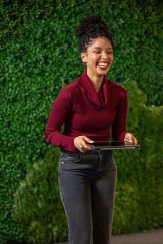 African american woman laughing while holding tablet in burgundy sweater and jeans by green foliage