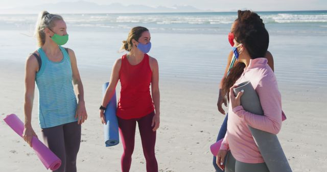 Group of friends wearing colorful face masks talking and holding yoga mats at a beach. Ideal for use in wellness blogs, fitness instructions, social distancing promotional materials, and outdoor activity advertisements. Represents healthy lifestyle and social wellness practices.