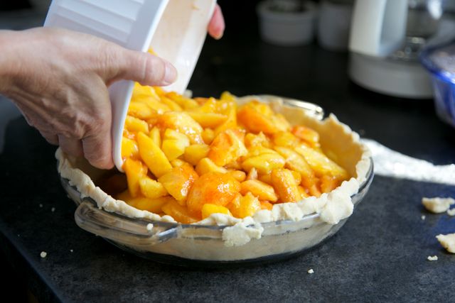 A close-up view of hands filling a homemade pie crust with fresh peach slices. The process takes place in a kitchen, indicating a home-cooked dessert preparation. This image is perfect for use in food blogs, recipe websites, cooking tutorials, and publications focused on homemade and rustic cooking.