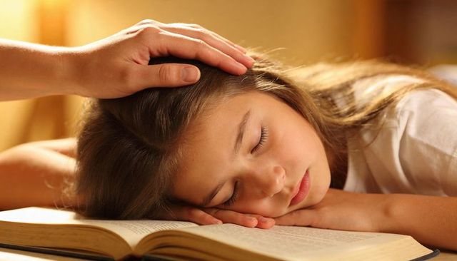 Young girl sleeping on open book while parent hand gently touching head, cozy study nap