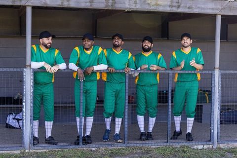 Five baseball teammates standing and cheering in dugout wearing green and gold uniforms
