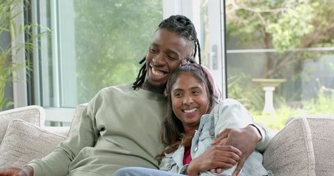 Diverse couple cuddling on couch at home, smiling and enjoying cozy daylight together