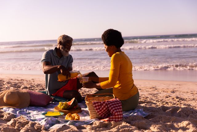 African american couple having food and drink while sitting on blanket against sea and sky in summer. picnic, copy space, nature, holiday, unaltered, love, togetherness, lifestyle and retirement.