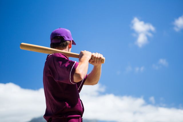 This image captures a baseball player in a purple uniform holding a bat, ready to play. The clear blue sky and sunny day create an ideal setting for outdoor sports. This image is perfect for use in sports-related content, advertisements, team promotions, or articles about baseball and outdoor activities.