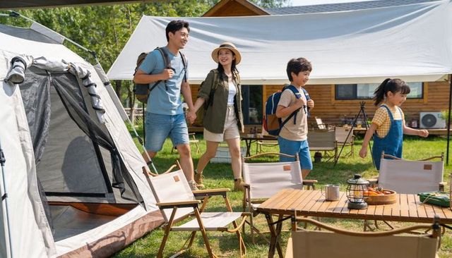Family of four walking to campsite carrying backpacks near cabin at sunny campground