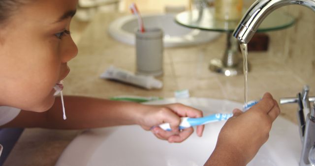 A young girl is intently watching as she rinses her toothbrush under running water in a bathroom sink, with copy space. Capturing a moment of daily hygiene routine, the image emphasizes the importance of teaching children about oral care.