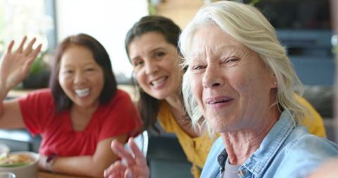 Smiling diverse women including senior friend waving and chatting at home dining table