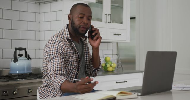 This image depicts a man sitting in a modern kitchen, multitasking by talking on the phone while writing in a notebook and working on a laptop. The kitchen is well-equipped with a blue kettle on stove. This image is useful for illustrating remote work, productivity, home office scenarios, or everyday multitasking.
