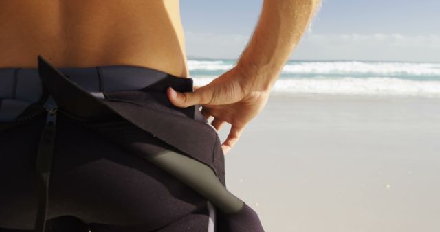 A person is adjusting their wetsuit at the beach, with copy space. Preparing for a surf session, the individual ensures their gear is secure against the backdrop of the ocean.