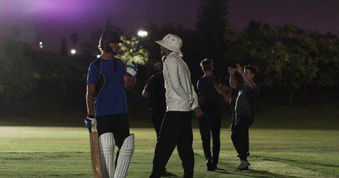 Night cricket practice showing batsman talking with coach under floodlights with helmet and pads