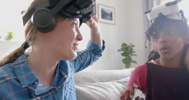 Two women sitting on a sofa, casually enjoying time together, wearing VR headsets. One woman is slightly lifting her headset while the other appears to look intrigued or engaged. Both are smiling, suggesting a pleasant experience. The scene is set in a well-lit living room with a houseplant and a picture frame in the background, creating a homely atmosphere. Ideal for technology, lifestyle, friendship, and indoor activity themes.