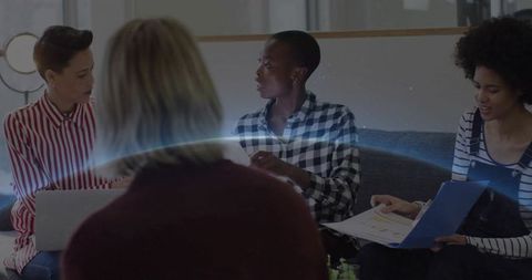 Diverse women collaborating in cozy lounge exchanging notes with laptop and blue folder