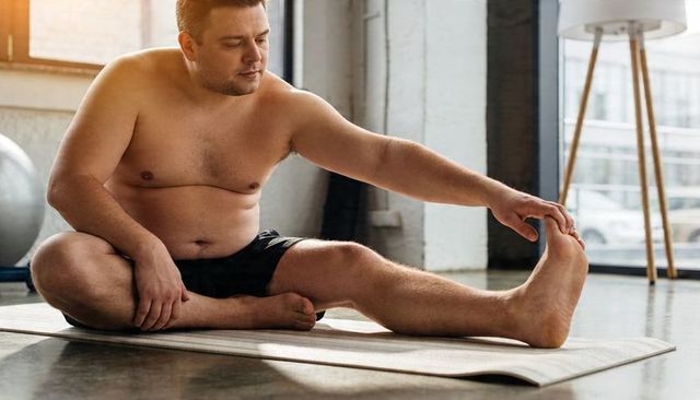 Plus-size man stretching on yoga mat in sunlit loft studio practicing flexibility