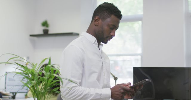 Young professional in white shirt using a tablet while standing in an office with window. Office environment includes desk, computer, and plant, creating a modern and fresh atmosphere. Ideal for business, technology, productivity, and modern workplace themes.