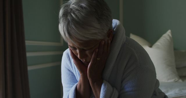 Senior woman holding her face in hands, feeling sad and stressed, sitting at home in a white robe. Mood appears contemplative and despondent. Useful for topics on mental health, aging, loneliness, senior care, and emotional wellbeing.