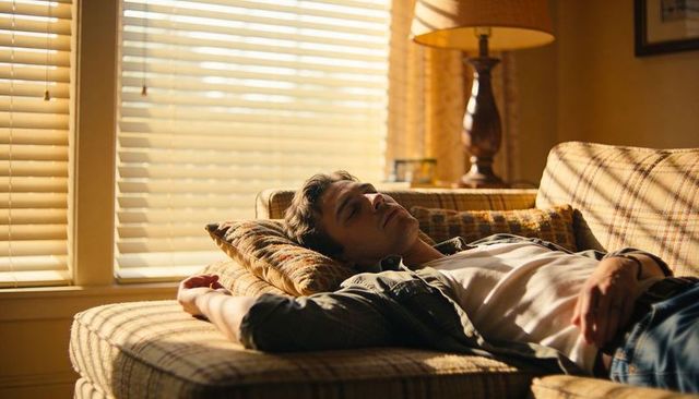Sunlit midcentury living room man reclining on plaid couch taking relaxed afternoon nap