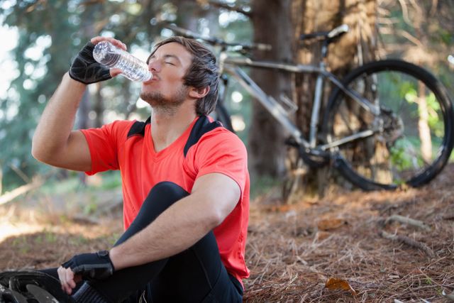 Male mountain biker taking a break and drinking water in a forest. Ideal for use in content related to outdoor activities, fitness, hydration, cycling, and healthy lifestyles. Perfect for promoting sports gear, hydration products, and adventure tourism.