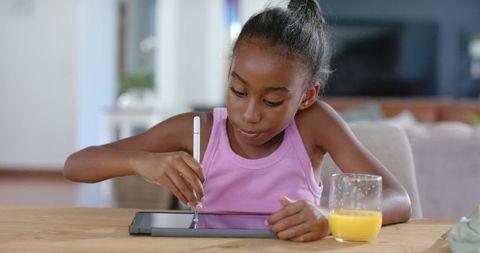 African american girl learning on tablet with stylus while enjoying orange juice at home