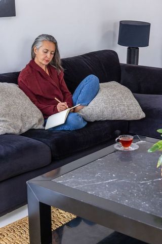 Senior woman writing in notebook on dark sofa wearing red cardigan with tea and plant