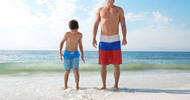 Father and son standing at the sandy beach, looking down at the waves. Both are wearing colorful swimwear, enjoying a sunny day by the ocean. Ideal for marketing summer vacations, family bonding themes, or promoting beachwear.