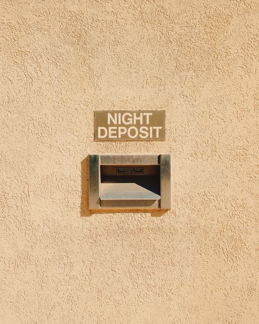 Image depicts a metal night deposit box mounted on a stucco wall with a sign overhead. This photo can be useful for illustrating banking services, security concepts, or financial institutions providing after-hours deposit services.
