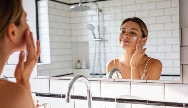 Smiling woman applying face cream at sink for morning skincare routine in tiled bathroom