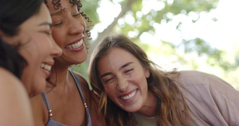 Laughing diverse female friends leaning heads together outdoors enjoying sunlit moment