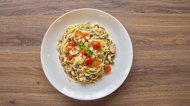 Plate of spaghetti mixed with sliced cherry tomatoes, mushrooms, and fresh basil served on a wooden table. Ideal for use in culinary websites, recipe blogs, and advertisements for Italian restaurants or healthy eating.