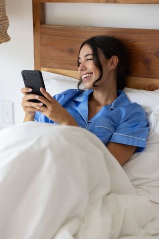 Smiling woman relaxing in bed with smartphone wearing blue pajamas cozy morning routine