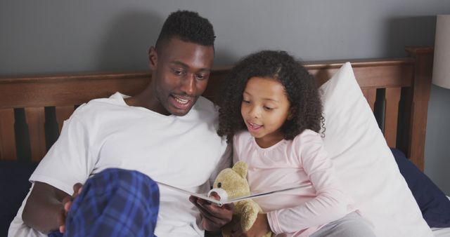 Happy african american father and daughter sitting up in bed reading story book. Fatherhood, childhood, care, togetherness and domestic life.