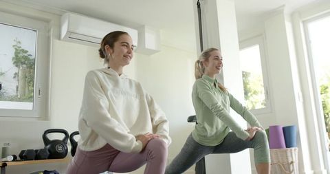 Two women doing lunges in bright minimalist home gym with kettlebells and yoga mats