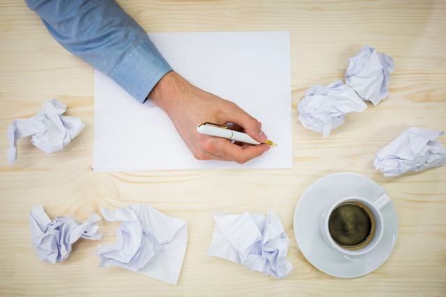 Businessman writing on blank paper surrounded by crumpled notes and a cup of coffee on a desk. Ideal for illustrating concepts of brainstorming, creativity, planning, and office work. Useful for business presentations, articles on productivity, and creative process visuals.