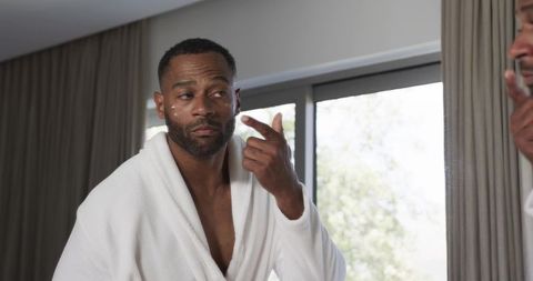 Man applying eye cream in white robe during morning grooming routine by window mirror