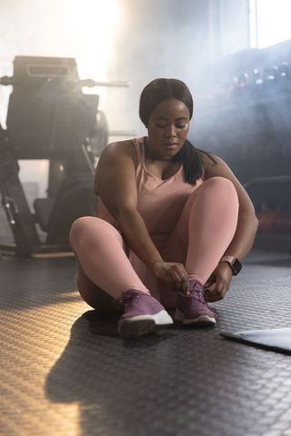 African american confident woman tying shoes in sunlit gym wearing pink leggings and smartwatch