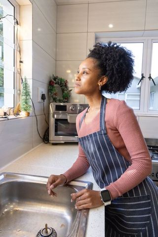 African american woman leaning at sink gazing out window wearing striped apron and smartwatch